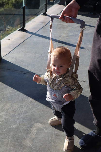 Toddler taking first steps in the Rest and Nurture baby walking harness while a parent holds the padded handle